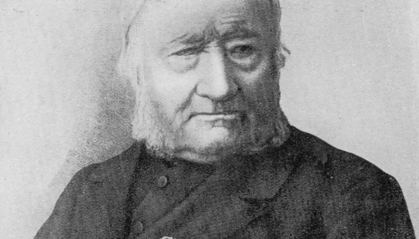 Portret Jana Dzierżona, pszczelarza z charakterystyczną brodą i siwymi włosami, w ciemnym stroju.