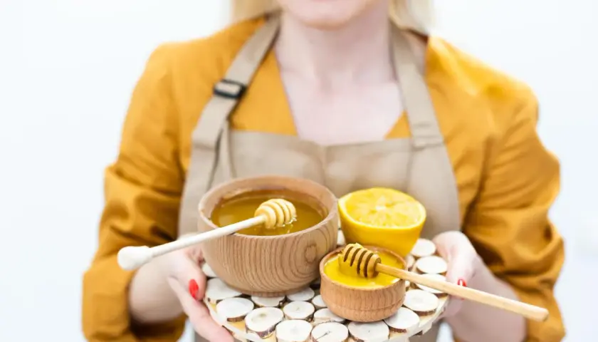 Kobieta w fartuchu prezentuje dwa drewniane naczynia z płynnym miodem i połówkę cytryny. Idealny zestaw na domowe SPA i naturalny vat na miód.