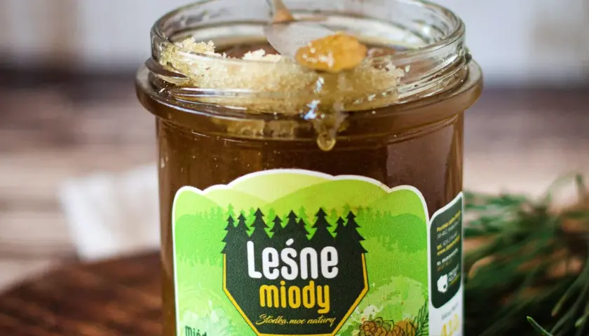 Słoik miodu iglasty, z kroplą spływającą z łyżeczki. Etykieta "Leśne miody" z sosnowymi drzewkami i szyszką.