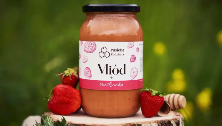 Słoik miodu truskawkowego z pasieki rodzinnej, otoczony świeżymi truskawkami i drewnianą chochlą.
